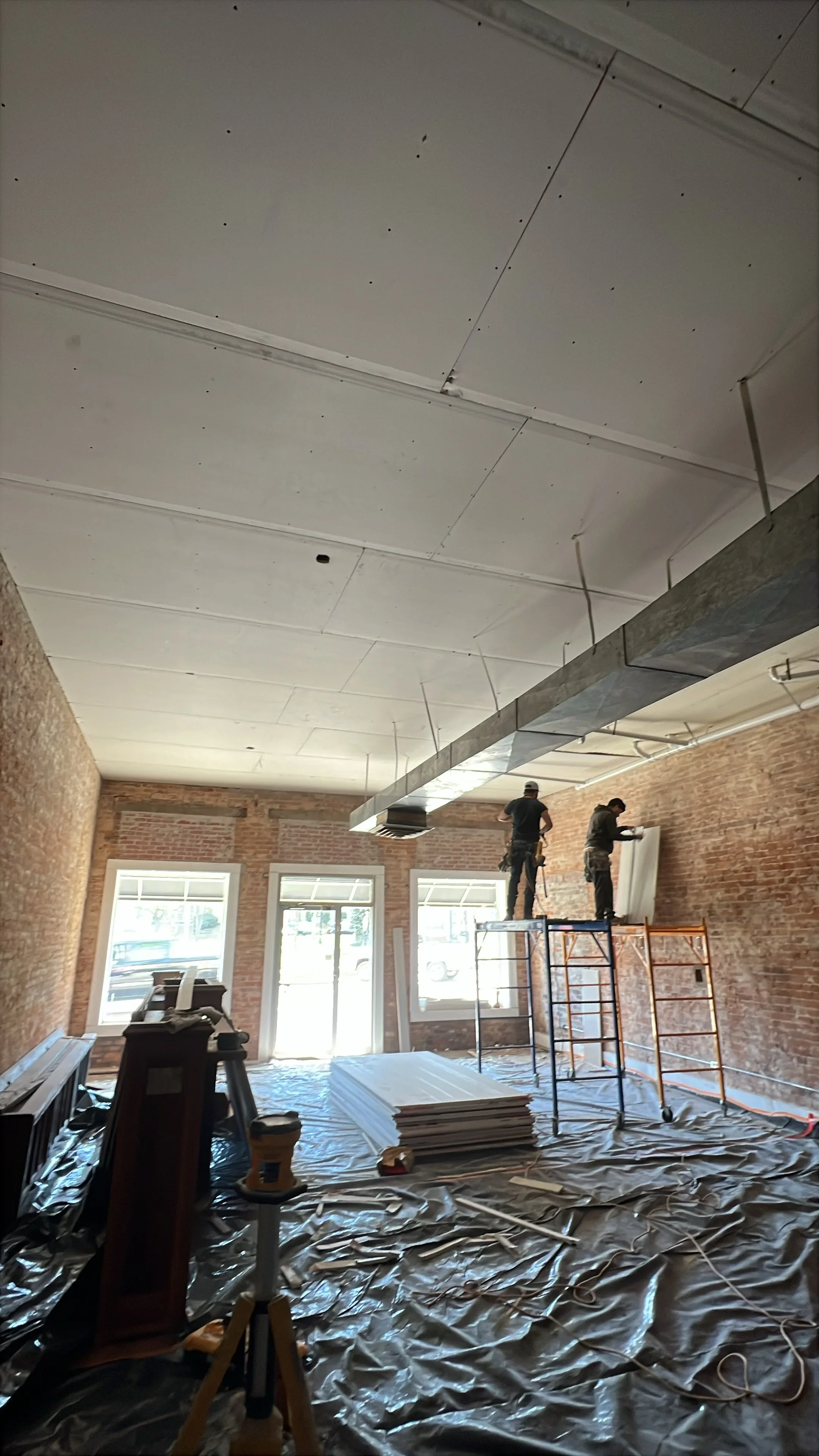 Commercial interior with white drywall ceiling, exposed brick walls, and workers on scaffolding during construction